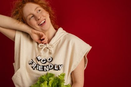 Cheerful woman with red hair in eco-friendly attire holding fresh lettuce against a vibrant red background.
