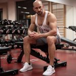 Strong, determined man in white tank top sitting in gym, showcasing fitness and strength.