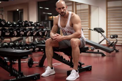 Strong, determined man in white tank top sitting in gym, showcasing fitness and strength.